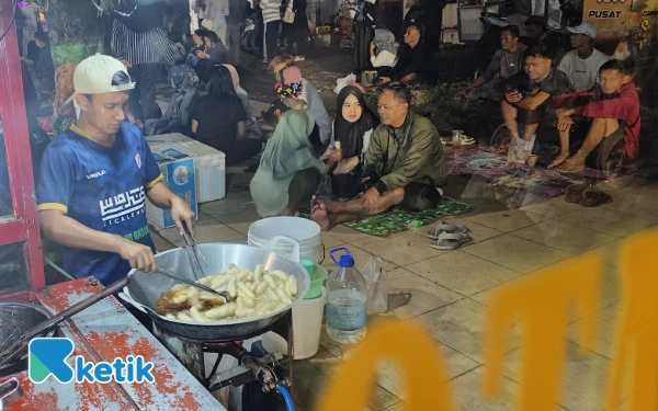 Foto Suasana ramai di sekitar gerobak gorengan milik Lakhood saat Festival Ronthek Pacitan 2025. Sambil menggoreng tahu dan otak-otak crispy, Lakhood melayani pengunjung yang duduk menikmati makanan di trotoar, menjadikan malam festival tak hanya meriah tapi juga menggerakkan ekonomi warga.