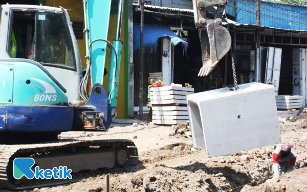 Foto Operator ekskavator mengangkat beton U-ditch dan pekerja menunggu di galian saluran untuk jalan beton. (Foto: Fathur Roziqq/Ketik.com)
